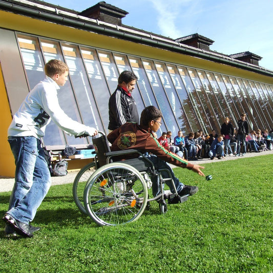 Ein Kind schiebt ein spielendes Kind im Rollstuhl während der Förderschullehrer daneben steht